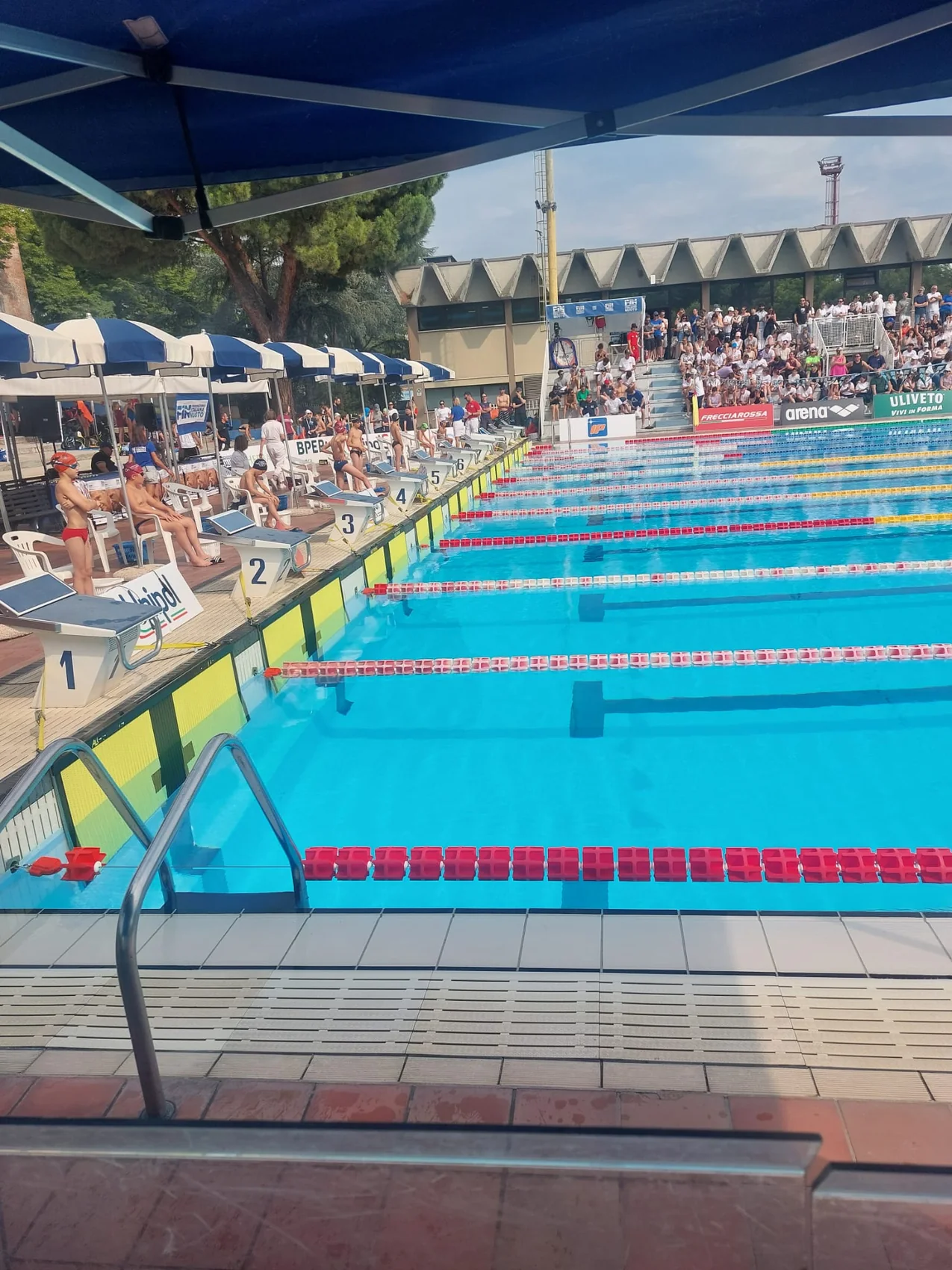 Piscina con blocchi di partenza, atleti e pubblico durante una manifestazione di nuoto.